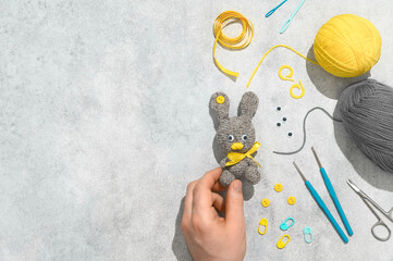 DIY Easter gift making theme. Male hand hold handmade knitted toy Easter rabbit and needlework accessories on grey background. Knitting concept. Overhead view, flat lay, copy space