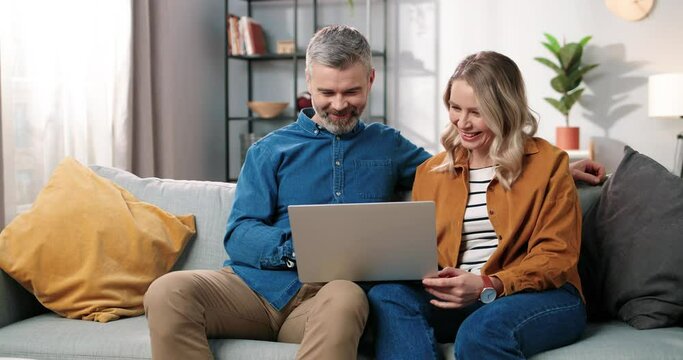 Caucasian middle-aged happy married couple bearded man and beautiful woman sitting on couch in good mood typing on laptop browsing online searching internet at home in cozy room, leisure concept