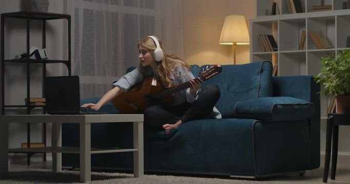 young woman is learning online playing guitar, viewing tutorial on laptop, hobby at free time at evening