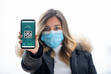 A defocused young girl wearing a face mask, is holding a smart phone with a digital illustration of a Covid-19 Health Passport concept.