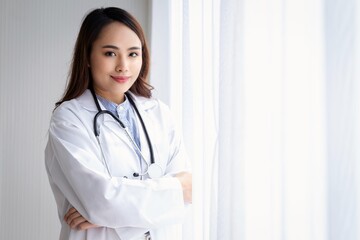 A female doctor in a white lab coat stands smiling by the window with white curtains in the sunlight.