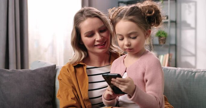 Close up portrait of happy Caucasian female parent mom sitting on couch in living room at home with little cute daughter tapping on cellphone surfing internet together, parenting concept