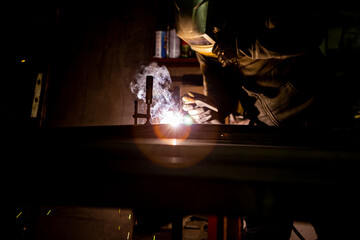 A welder welds metal pipes in an old workshop. The worker has a protective welding mask on his head. Copy Space.