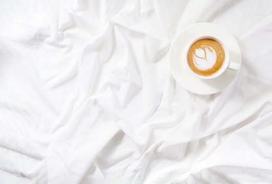 Cup of coffee cappuccino in bed on a white sheet of bedding linen. Cozy soft flatlay morning routine. Copy space mockup. Concept minimalism.