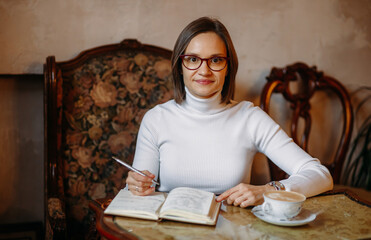 Fototapeta na wymiar Young business woman in glasses in a white sweater sits in coffee at the table holds a pen and writes looks at the camera