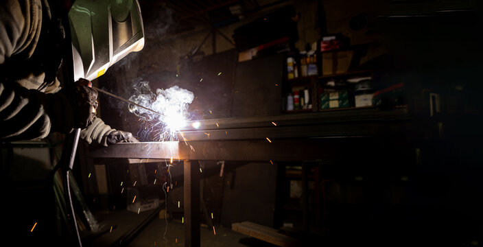 A man with a protective mask on his head welds pipes in a workshop. Copy space.