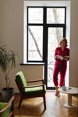 Full length view of smiling european woman reading magazing in morning. Indoor shot of lovely barefoot girl in pajama standing near window.