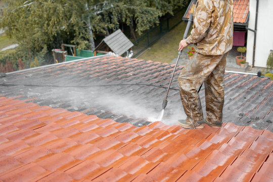 Professional In Uniform Using Special Equipment To Remove Dirt From Rooftop. In The Background Visible Children Playground And Backyard. Before And After Washing State Of Roof. Cleaning Service.