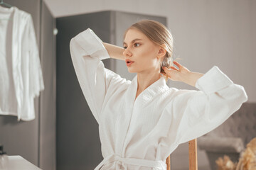 Young blonde beautiful woman in the white bathrobe looking to the mirror and touching her hair