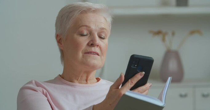 Old Caucasian woman gray-haired elderly mature lady sitting at home reading book refuses literature for electronic device phone looks at smartphone screen straightens her hair, technology concept