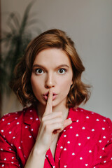 Fototapeta premium Front view of gorgeous woman showing hush sign. Indoor shot of curly female model in red pajama.