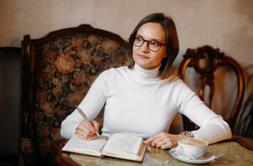 Fototapeta na wymiar Portrait of a young business woman in glasses sitting in a cafe with a pen in her hands and looking to the side