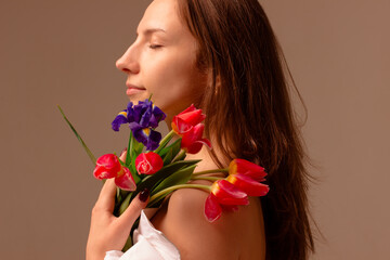 Fototapeta premium portrait of a sensual girl in profile with flowing hair and bare shoulders closed her eyes stands and dreams holding flowers, on the shoulder on the background beige background