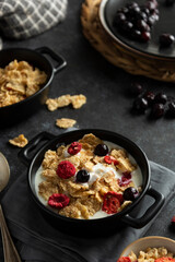Corn flakes with strawberry and blueberry in black bowls over dark background, top view.