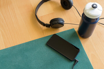 Green mat, headphones, smartphone and blue bottle of water on wooden floor. Concept of physical activity lessons at home via the internet, with copy space.