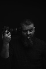 Portrait of a man with a beard, holding a camera in his right hand, pointing to his temple and showing his tongue, portrait of a man on a black background, soft focus, artistic noise