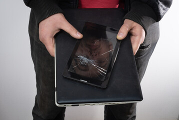 A broken smartphone with a cracked screen and a face reflection on it, and a broken laptop in the hands of a young man.