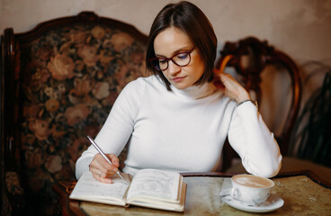 Smart young woman with glasses sits at a table in coffee and ponders the data in a notebook. Looks at notepad