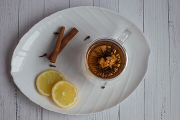 The tea flower is brewed, opened in a transparent cup on a white platter, with cinnamon and cloves
