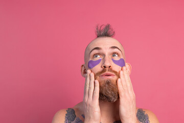 European handsome bearded tattooed topless man with purple eye patches mask posing to camera, face care for men. Isolated on pink background.