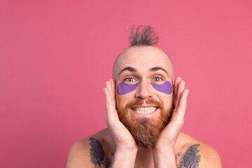 European handsome bearded tattooed topless man with purple eye patches mask posing to camera, face care for men. Isolated on pink background.
