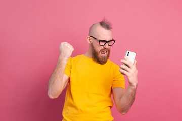 Image of screaming young bearded handsome man standing isolated over pink wall background using mobile phone make winner gesture