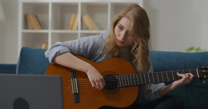 pretty young woman picking up tune on guitar, sitting alone at living room, spending time at home at evening