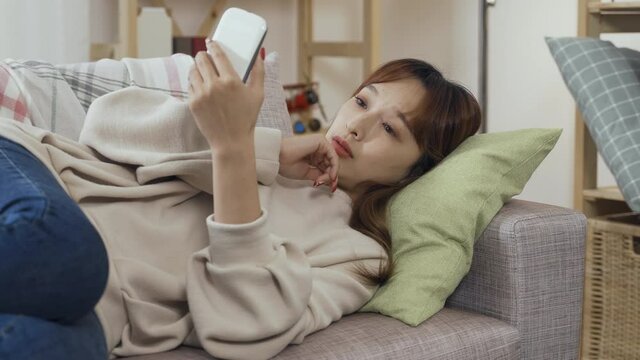 lazy ambience korean girl reclining on sofa phubbing smartphone feels bored and yawns. spiritless asian woman doing nothing but using her phone in living room didn't see anything interesting.