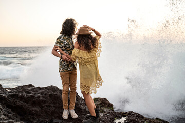 Back view of Happy young hippie couple having fun at the beach, looking at big waves.