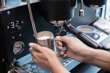 Closed up of barista’s hand holding pitcher full of milk frothing from coffee machine. For making cappuccino or latte