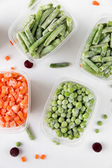 Frozen vegetables in transparent containers on a white background stand on top of each other, next to pieces of vegetables, vertical photo