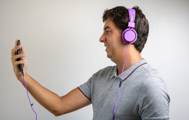 Brunette man listening to music at home with purple headphones and a cellphone. Wearing a gray polo...