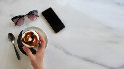 Girl's hand keeps a glass with freshly made testy coffee drink with whipped cream and syrup next to fashionable sunglasses and smartphone on the marble table. Copy space