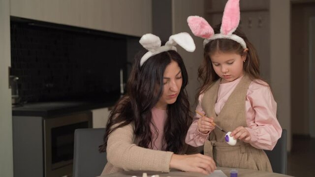 A cute family mom and daughter are preparing for Easter by painting chicken eggs with a brush. The girl draws circles on a white egg.