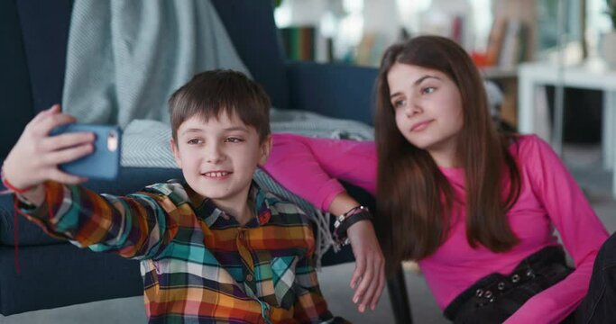 Funny adorable teen couple of elder sister and young brother using mobile phone making online video broadcast blog for followers on social media. Siblings.