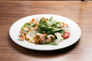 Caesar salad with prawns, lettuce, quail eggs, cherry tomatoes, croutons and parmesan cheese in a white ceramic plate on the wooden kitchen table.