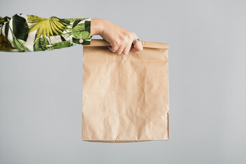 Female hand holds a paper bag against gray backdrop. Empty eco friendly package, gift, surprise or delivery concept - obrazy, fototapety, plakaty