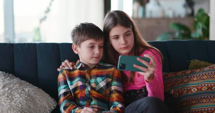 Adorable couple of teenage siblings using mobile phone watching favorite funny videos on social media application browsing technology staying home.