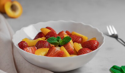 Light simple fresh berry and fruit salad in a white plate with mint. Selective focus on mint plate. Concept of low calorie delicious breakfasts and snacks. Grey background.