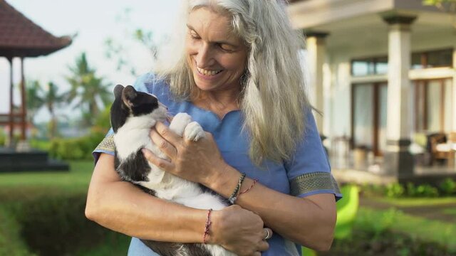 Happy senior grey-haired woman holds a cat in her hands and strokes and caresses a pet outdoor