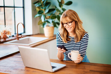 Mature woman using her mobile phone and laptop while sitting in her kitchen at home