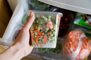 Different frozen vegetables in a transparent box is held by a female hand, in the background a freezer with vegetables, life style, close-up