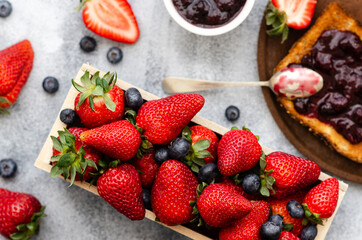 Strawberries and blueberries in a wooden rectangular box, a slice of toast with strawberry and blueberry jam on a wooden plate and a spoon, and strawberry and blueberry jam in a white bowl. Top view. 
