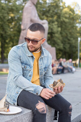 Hipster man eating burger on the street. Young bearded stylish guy biting delicious burger.Close up of young man eating burger outdoors. Man with beard having street food.