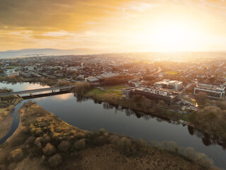 Fototapeta premium Aerial drone view on Sunset over Galway city, Bridge over River Corrib and NUI buildings, Atlantic ocean in the background. West coast of Ireland. Warm sunny cloudy sky, Sun flare