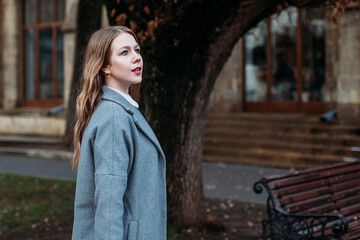 European young woman in coat walks in autumn park, enjoy autumn time outside. Beautiful yellow foliage on bushes and trees. Autumn mood. Fall season..
