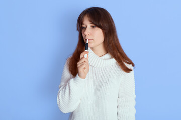 Attractive European young woman smoking electronic cigarette indoor isolated over blue background, looking away, wearing casual white jumper, has long dark hair.