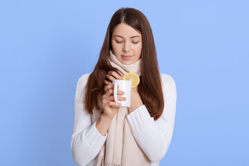 Woman warming up with cup of tea, looking down, keeping hand on neck, has sore throat, wrapped in scarf, wearing white scarf, lady with pleasant appearance enjoying hot beverage against blue wall.