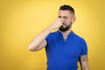 Handsome man with beard wearing blue polo shirt over yellow background smelling something stinky and disgusting, intolerable smell, holding breath with fingers on nose