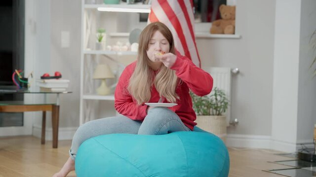 Relaxed Korean teen girl watching TV and eating pizza sitting on bag chair indoors. Wide shot portrait of Asian teenager enjoying leisure at home with American flag at background. Adolescence.